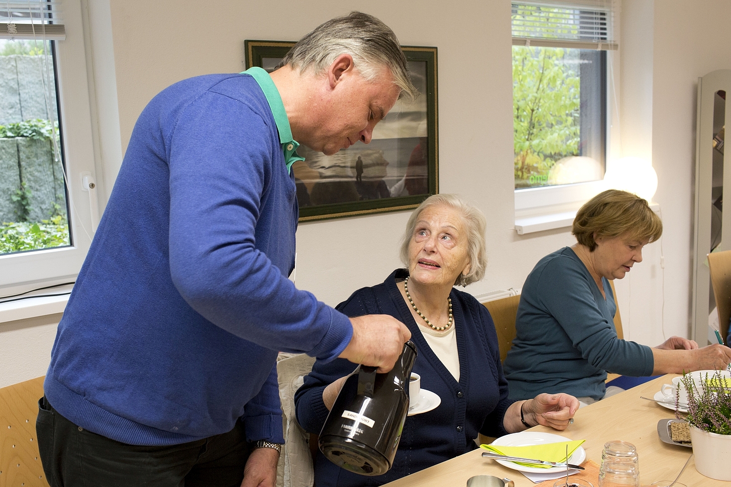 Foto: Mann schenkt älterer Frau Kaffee ein, eine weitere Frau sitzt daneben
