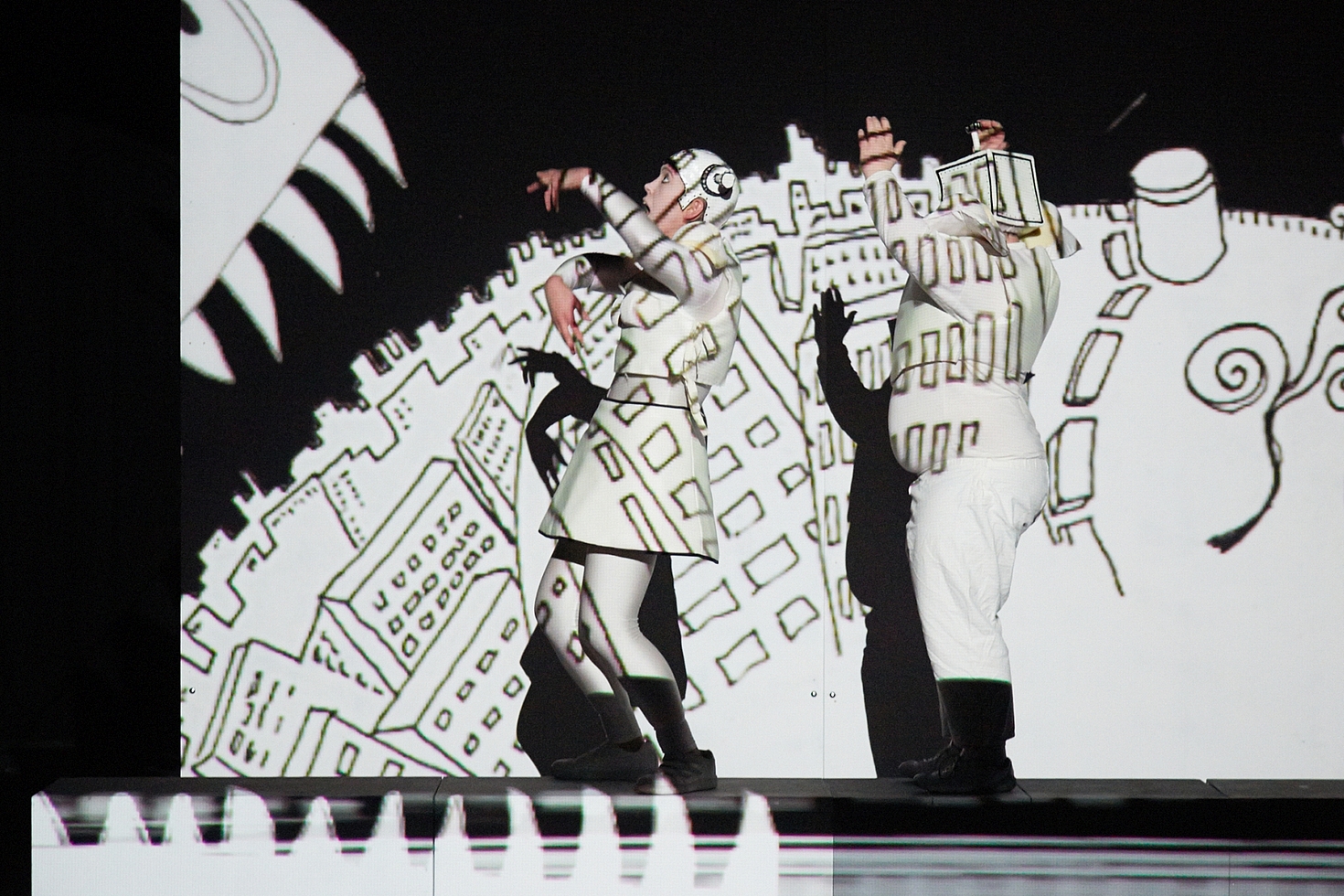 Foto: Zwei Schauspieler vor einer Bühnenwand mit Diaprojektion einer comichaften Stadt